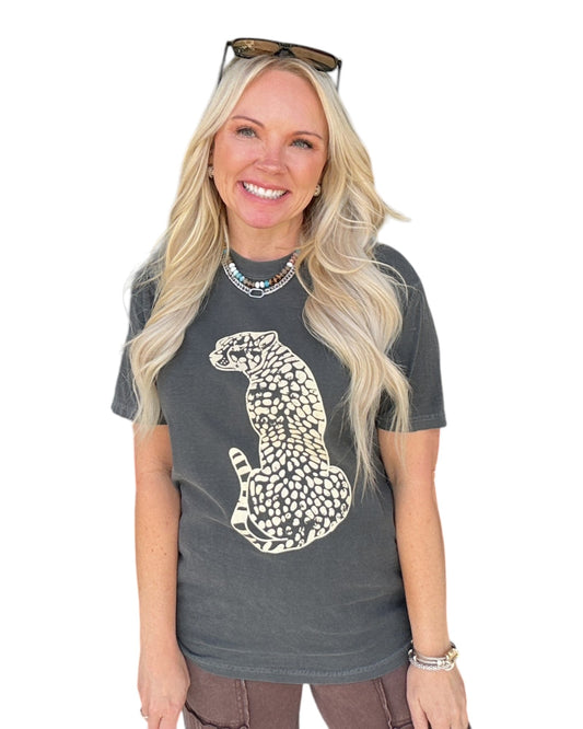 A smiling blonde woman wearing a dark charcoal gray t-shirt with a cream-colored spotted cheetah graphic on the front, styled with sunglasses on her head and a beaded necklace, against a plain white background.